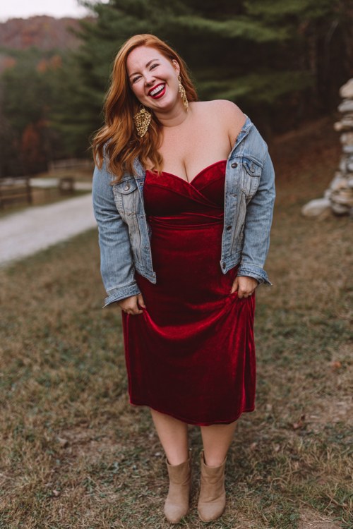 A curvy woman wears a deep red velvet dress with a sweetheart neckline, styled with a light-wash jean jacket and ankle boots