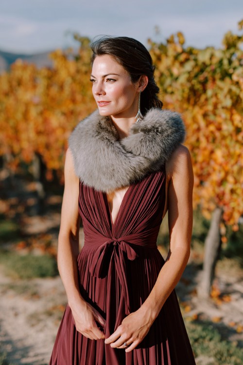 A woman wears a maroon pleated maxi dress styled with a light gray fur stole, standing by a vineyard ceremony with orange and gold leaves in the background