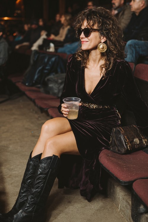 A woman over 40 wears a dark velvet wrap dress with a side slit, paired with tall black cowboy boots and gold hoop earrings