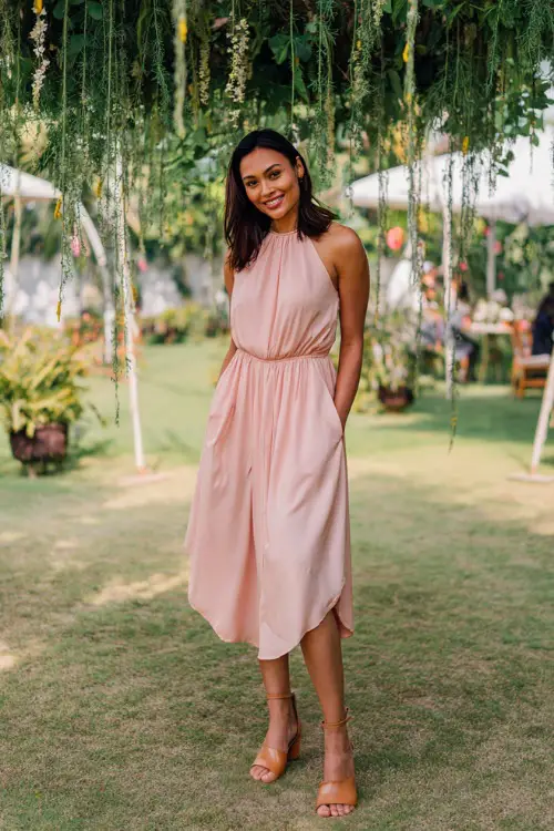 A woman wearing a soft pink sleeveless midi dress with gathered waist and tan block heels, posing under a canopy of hanging greenery at an outdoor wedding