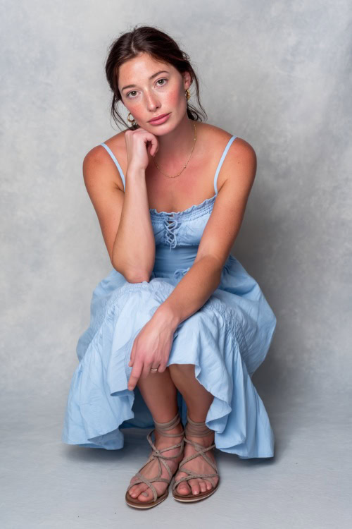 A woman in a light blue smocked sundress with simple jewelry and flat lace-up sandals, styled for a soft casual evening look, simple studio background