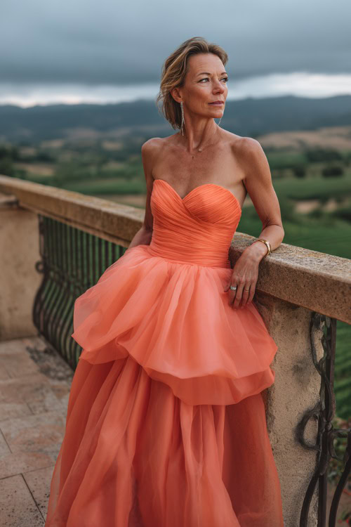 A woman over 50 wearing a coral A-line gown with a structured bodice and layered tulle skirt, posing on a balcony overlooking a vineyard wedding setting