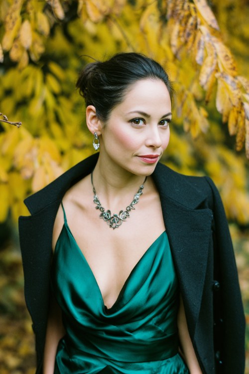 A woman wears a deep emerald green satin gown with a draped neckline, styled with a black tailored blazer and statement necklace