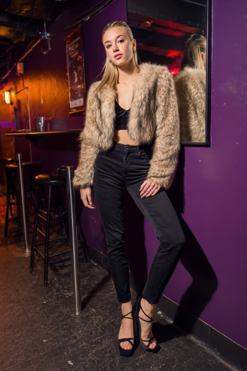 A woman in skinny black jeans, a faux fur jacket, and strappy heels, posing inside a nightclub with purple lighting