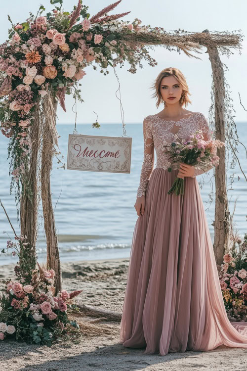 A woman in a dusty rose A-line dress with lace cap sleeves, standing beside a beachside wedding welcome sign adorned with delicate spring florals