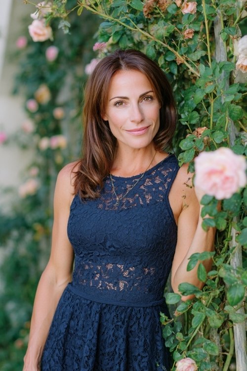 a woman over 50 in a navy fit-and-flare dress with lace overlay, posing near a trellis wrapped in climbing roses