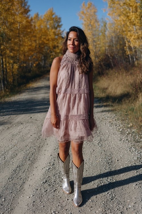 A woman over 30 wearing a blush pink A-line dress with a tiered sheer skirt and high neck, paired with metallic silver cowboy boots