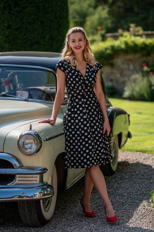 A woman in a black and white polka dot cocktail dress with cap sleeves and red heels, smiling beside a classic vintage car parked at a garden wedding