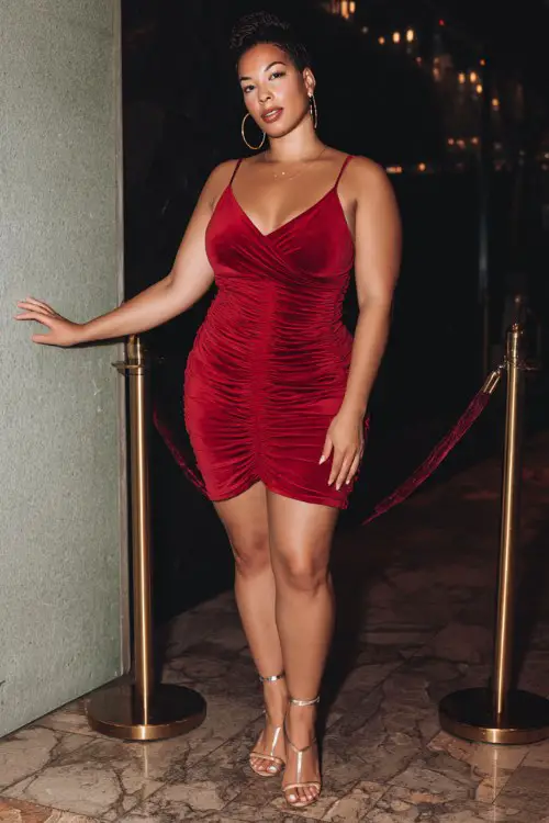 A curvy woman wears a red ruched bodycon dress with strappy stiletto heels and gold hoop earrings