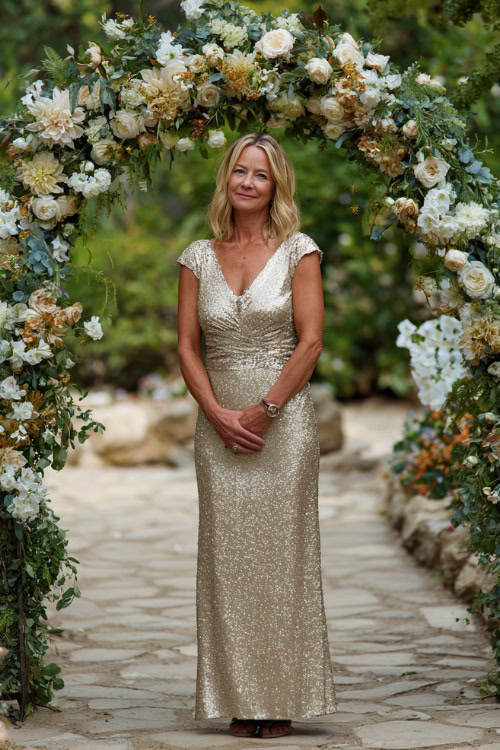 A woman over 50 in a champagne-colored sequin dress with cap sleeves and a V-neckline, standing under a wedding arch covered in summer florals