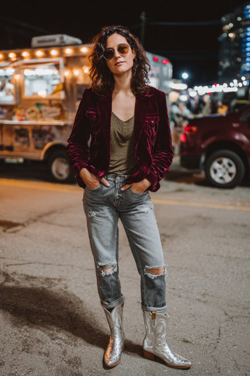 A woman over 30 wears distressed jeans, a velvet burgundy jacket over a fitted band tee, and metallic silver cowboy boots