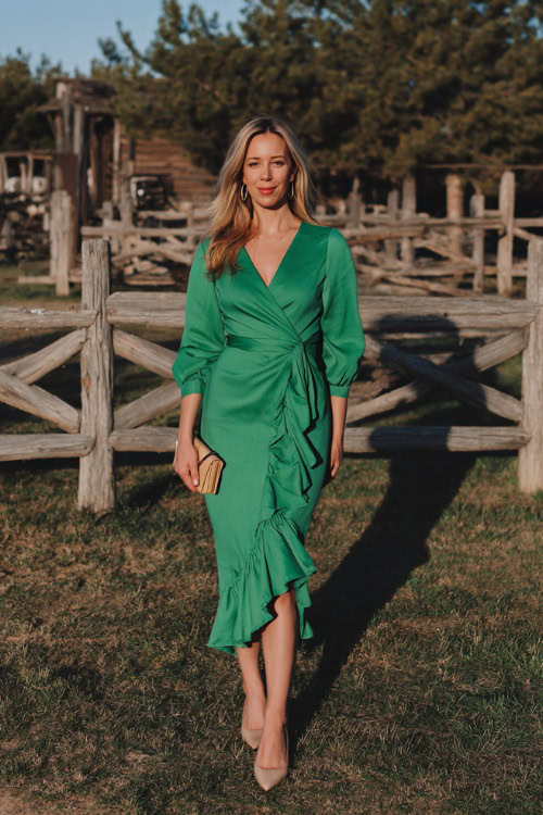 A woman wears a bright pine green midi wrap dress with a ruffled hem, styled with beige pumps and a small clutch