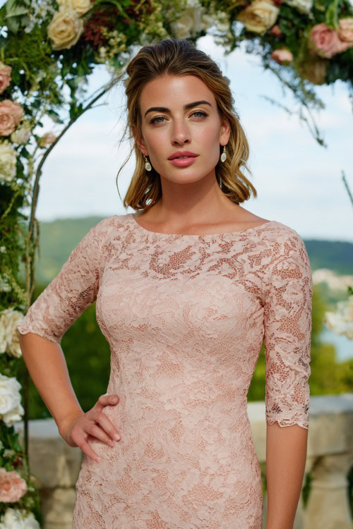 A woman wearing a blush pink lace sheath dress with elbow-length sleeves and drop earrings, standing beneath a floral arch at a formal outdoor wedding
