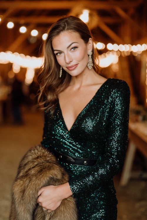 A sparkling sequin wrap dress in deep emerald green with a side slit, styled with black leather cowboy boots and a faux fur stole for warmth