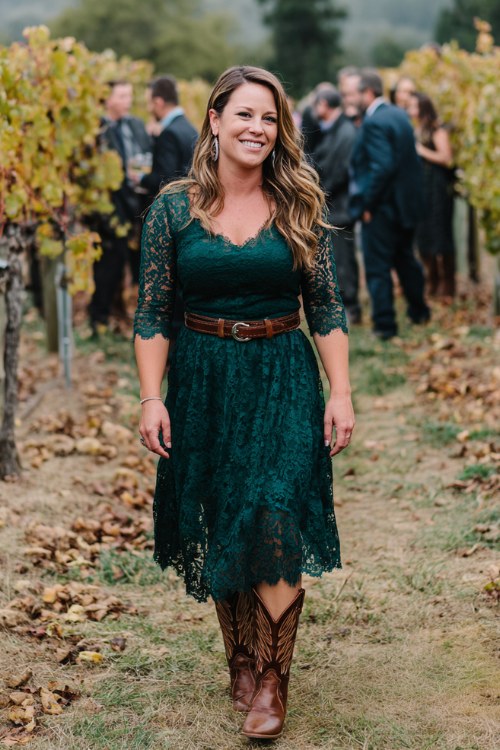A curvy woman over 40 wears a forest green lace midi dress with sheer sleeves and dark tan cowboy boots, styled with a leather belt