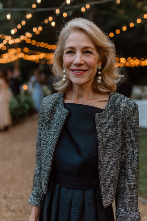 A woman over 50 wears a navy long-sleeve gown with a tweed blazer and pearl earrings