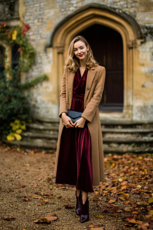A woman wears a burgundy velvet midi dress with long sleeves, layered under a tailored camel wool coat