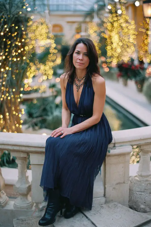 A woman over 50 wears a dark blue pleated chiffon gown with a beaded neckline and heeled boots