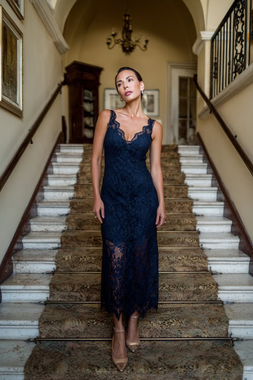 A woman over 50 wearing a navy lace overlay gown with scalloped edges and low heels, standing at the top of a staircase inside a classic estate wedding venue