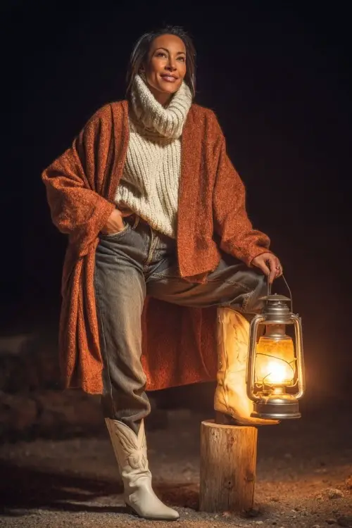 A woman over 40 in high-waisted jeans, a knit turtleneck sweater, and white cowboy boots with a wool wrap coat near glowing lanterns