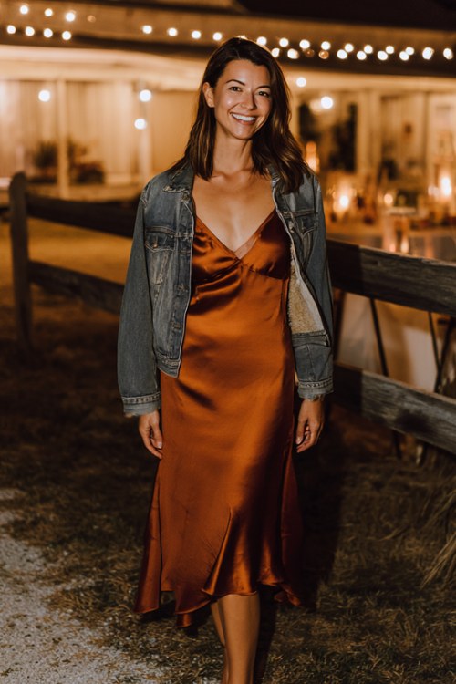 A woman wears a copper satin slip dress with a flowing hem, paired with a fitted jean jacket and strappy heels