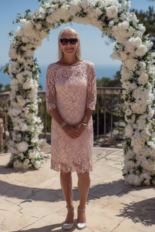 A woman over 60 in a soft blush pink A-line dress with lace overlay and block heels, posing beside a white floral arch at an outdoor ceremony, full body view