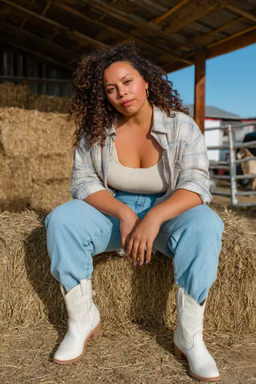 A curvy woman wears light blue straight-leg jeans, white cowboy boots, and a loose cream flannel layered over a ribbed tank top