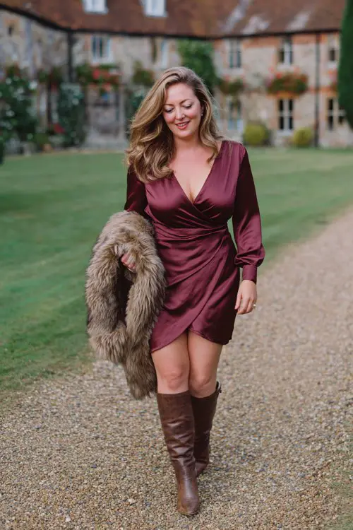 A curvy woman wears a burgundy satin wrap dress with long sleeves, styled with brown leather knee-high boots and a faux fur stole