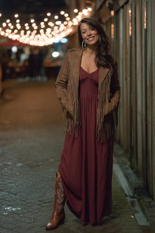 A woman over 40 in a burgundy maxi dress with a suede fringe jacket and tall cowboy boots, accessorized with hoop earrings under glowing string lights