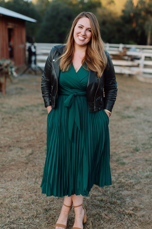 A curvy woman wears a forest green pleated midi dress with a tie waist, paired with a cropped leather jacket
