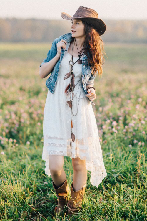 A woman wears cowboy boots with a long white dress and a denim coat