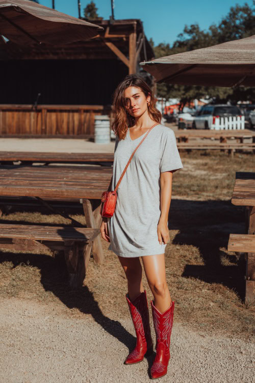 A woman wearing a casual T-shirt dress in light grey, paired with red western boots and a small crossbody bag