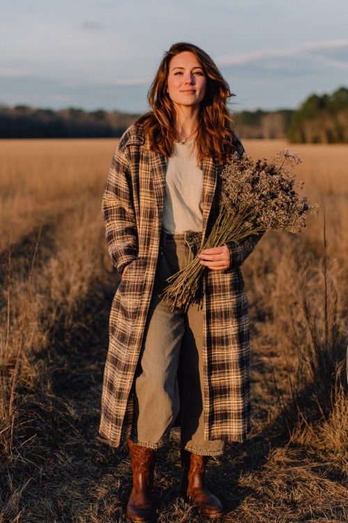 A plus-size woman wears wide-leg jeans, a long plaid duster coat over a simple tee, and dark brown western boots