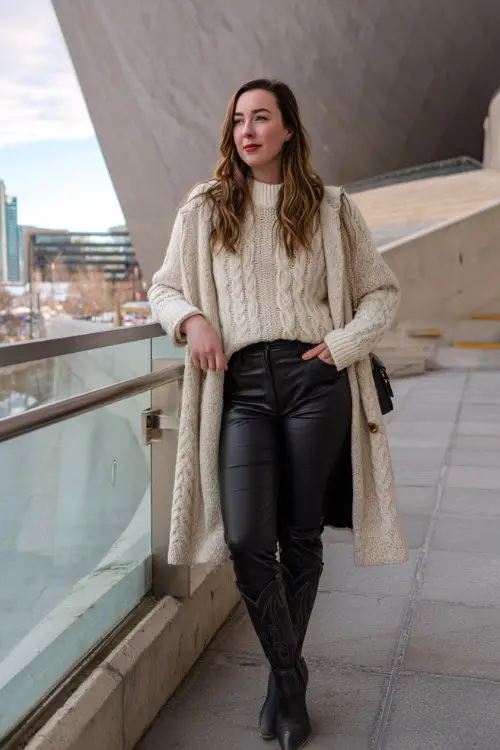 A curvy woman wears black cowboy boots with faux leather leggings, a cream cable-knit sweater, and a structured wool coat