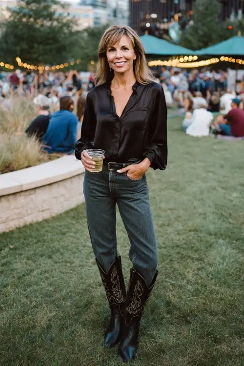 A woman over 40 wears bootcut jeans with a silk blouse tucked in and sleek black cowboy boots