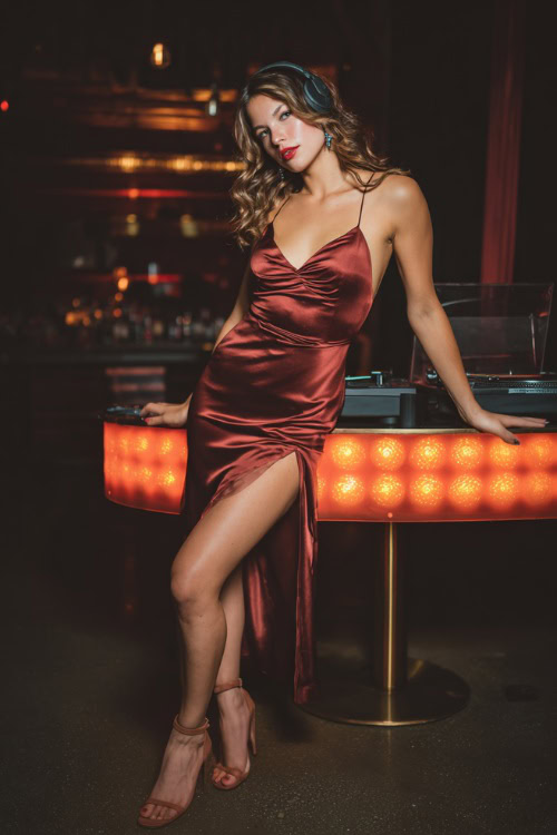 A woman in a fitted burgundy satin slip dress with a thigh slit, paired with ankle strap heels, posing near a glowing DJ booth in a fall nightclub