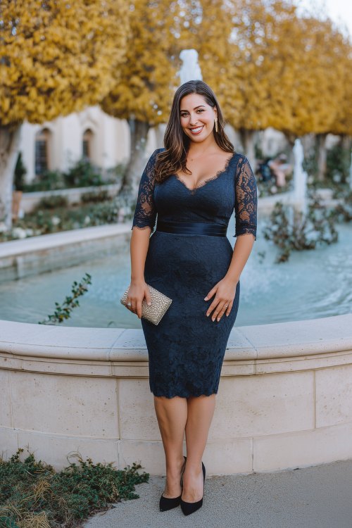 A curvy woman in a navy sheath dress with lace sleeves and a knee-length hem, paired with black slingback pumps and a pearl-embellished clutch