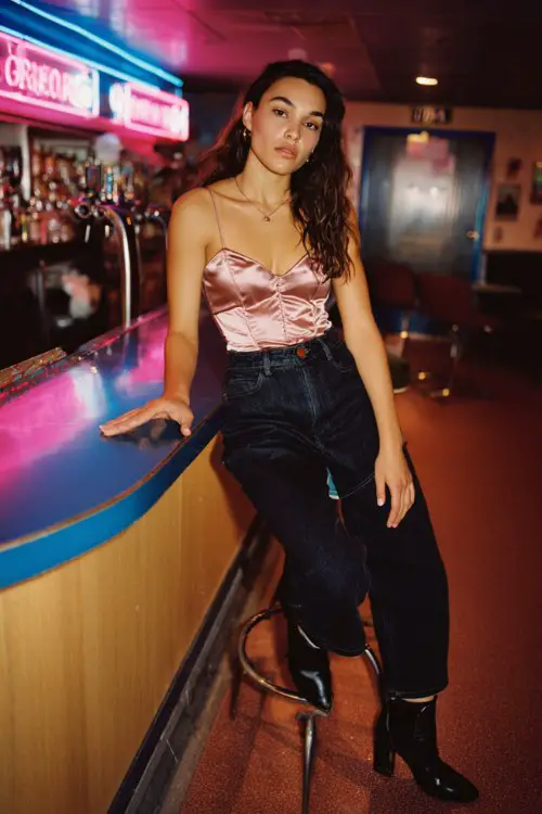A woman wears dark denim jeans with a corset-style satin top and heeled ankle boots, leaning on a bar counter