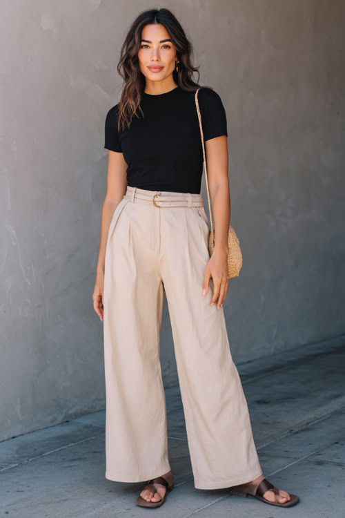 A woman in a basic black tee tucked into beige wide-leg pants with flat mules and a straw crossbody bag, styled for casual city nights, simple studio background