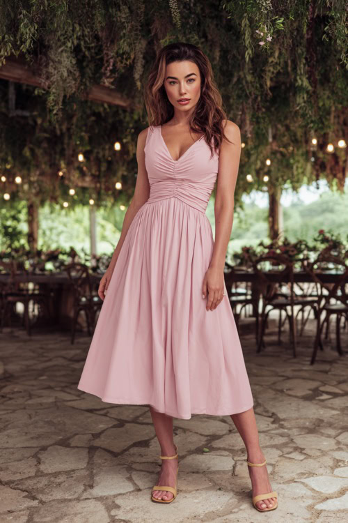A woman wearing a soft pink sleeveless midi dress with gathered waist and tan block heels, posing under a canopy of hanging greenery at an outdoor wedding