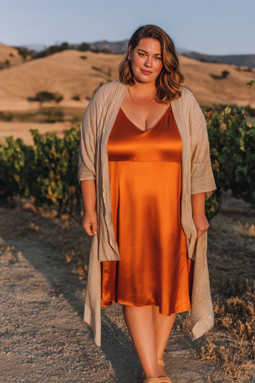 A plus-size woman wears a rust-orange satin slip dress layered with a light shawl, styled with gold sandals and minimal jewelry