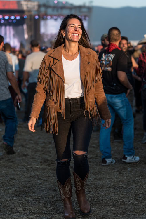 A woman over 30 wears ripped black jeans with a suede fringe jacket, paired with a white tee and western boots, standing in a lively country concert crowd