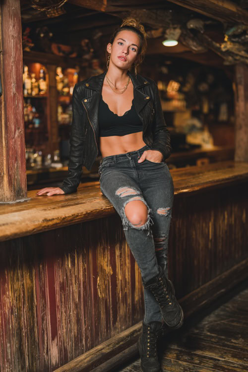 A woman in ripped skinny jeans, a black cropped leather jacket, and lace-up boots, leaning against a rustic wooden bar on a fall night, full body outfit visible