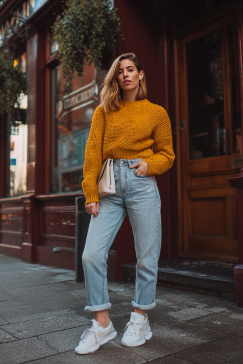 A woman wearing mom jeans, a mustard yellow knit sweater, and white low-top sneakers, carrying a small crossbody bag outside a neighborhood pub