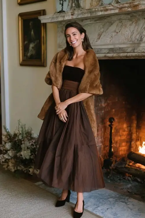 A woman wears a chocolate-brown organza gown with layered skirt and velvet waistband, layered with a camel wool cape coat and suede ankle-strap heels