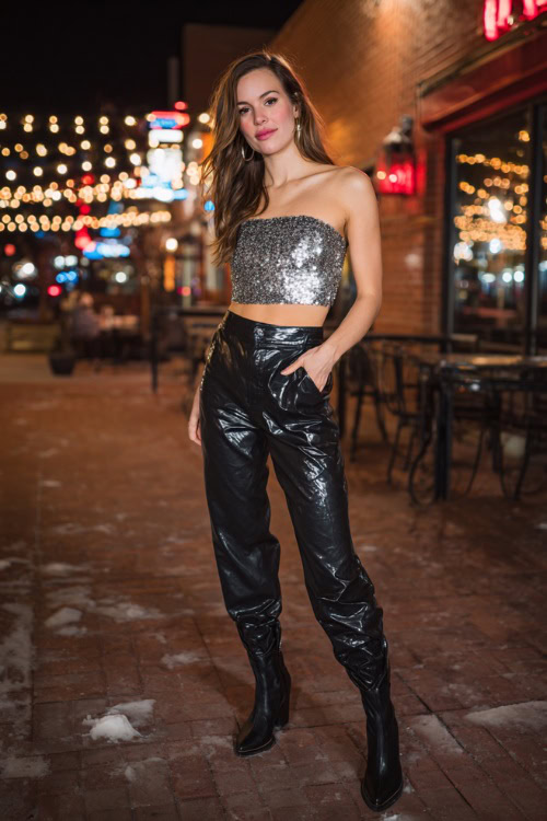 A woman wearing black faux leather pants, a sequined silver crop top, and heeled ankle boots, posing at the entrance of a busy casual bar on a crisp fall night