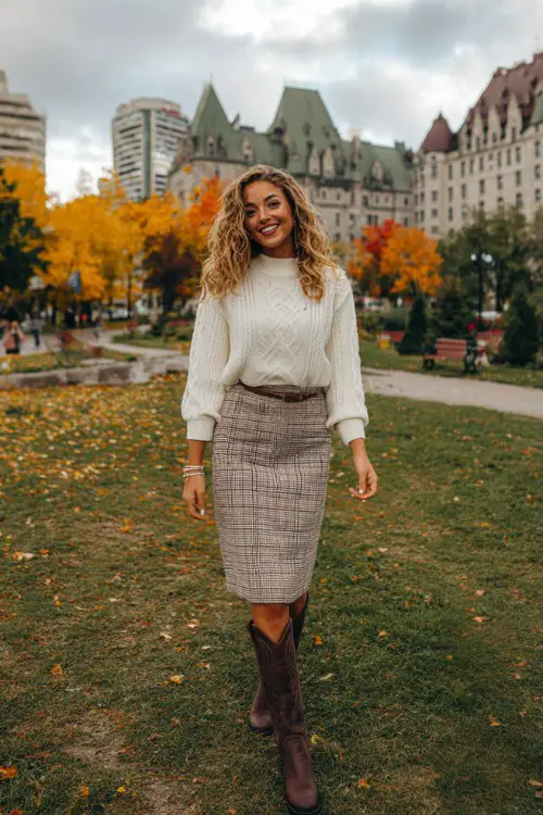 A woman over 30 wears brown tall cowboy boots with a cream sweater and plaid skirt