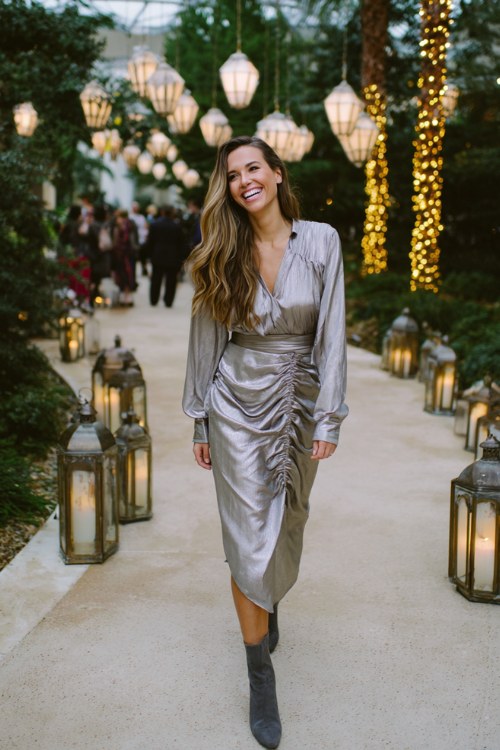 A woman wears a silver metallic long-sleeve midi dress with subtle shimmer and sculptural draping, paired with dark gray suede booties