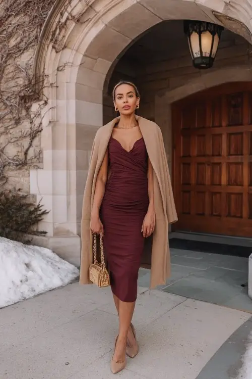 A woman wears a burgundy wrap midi dress with side ruching, styled with a camel wool cape coat, pointed-toe pumps, and a gold chain bag