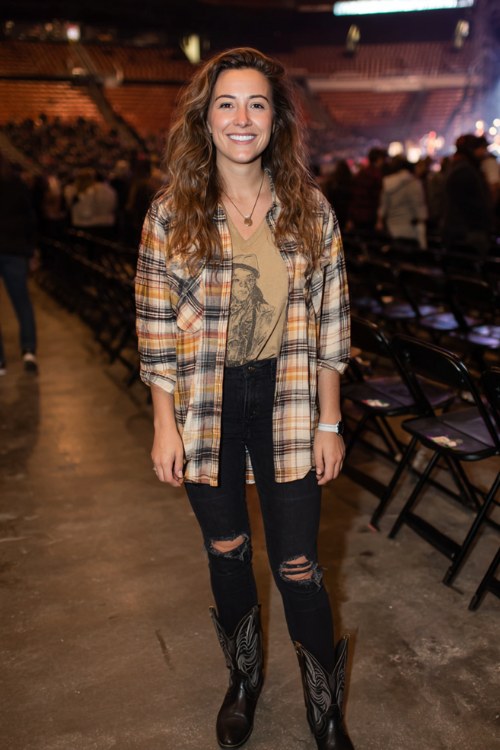 A curvy woman wears ripped black jeans with a graphic tee layered under a plaid overshirt, styled with black cowboy boots